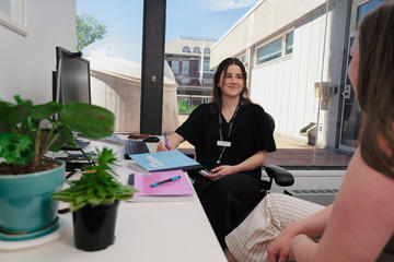 Staff member assisting a visitor at a desk beside a window, with notebooks, computer equipment, and potted plants.