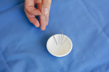 Hand holding an acupuncture needle above a dish containing several more needles on blue fabric.